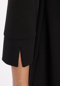 Robe noire avec un tissu texturé, des manches trois-quarts et un détail fendu au niveau du poignet. Design simple et épuré.