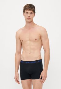 Junger Mann mit kurzen braunen Haaren, der schwarze Boxershorts von Jack & Jones trägt und vor einem schlichten weißen Hintergrund steht.