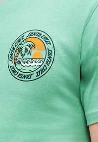 Κυκλικό λογότυπο σε ανοιχτοπράσινο πουκάμισο με φοίνικα, κύματα ωκεανού, δύση του ήλιου και το κείμενο «Santa Cruz» επαναλαμβανόμενο γύρω από τον κύκλο.