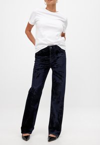 Mustat samettiset leveälahkeiset housut, joissa on hienovarainen kiilto, yhdistettynä yksinkertaiseen valkoiseen t-paitaan ja mustiin suippokärkisiin korkokenkiin. Näkyvä sileä tekstuuri.