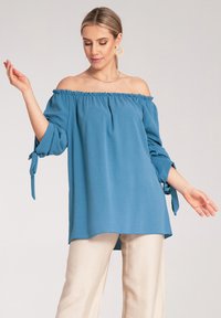 Jasnoniebieska bluzka bez ramion z elastycznym dekoltem, długimi rękawami z wiązanymi akcentami i luźnym krojem, który miękko układa się na sylwetce.