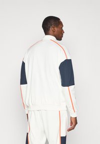 Λευκή αθλητική ζακέτα με ναυτικές μπλε λεπτομέρειες και πορτοκαλί ραφές. Διαθέτει ψηλό γιακά και πλεκτούς μανσέτες για άνετη εφαρμογή. Λείος υφασμάτινος ιστός.