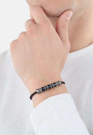 Pulsera de cordón negro trenzado que presenta tres acentos metálicos cilíndricos con ranuras y detalles suaves en azul, llevada en una muñeca.