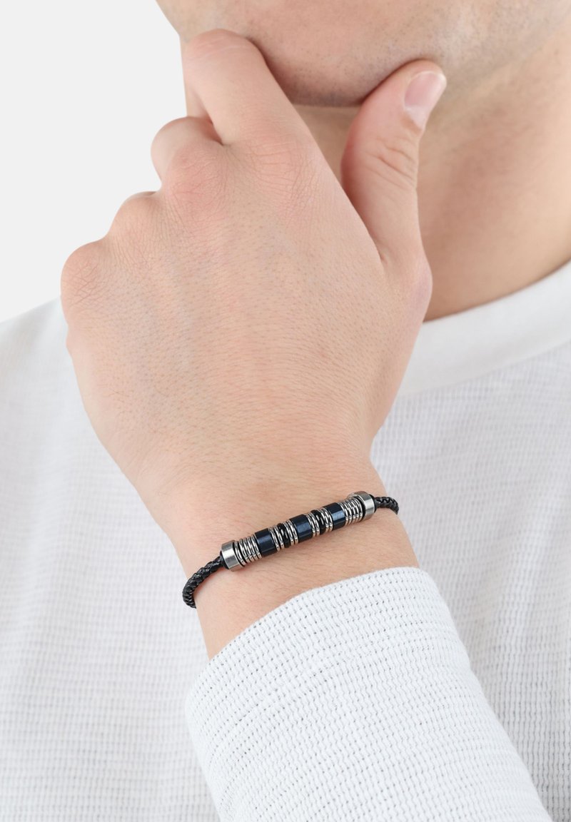 Pulsera de cordón negro trenzado que presenta tres acentos metálicos cilíndricos con ranuras y detalles suaves en azul, llevada en una muñeca.
