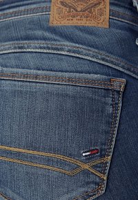 Denimjeans i en mörkblå nyans, med kontrasterande orange sömmar, en läderlapp och en liten broderad logotyp i röd, vit och blå.