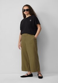 Zwarte t-shirt met korte mouwen en een klein wit grafisch ontwerp, gecombineerd met losse olijfgroene wijde broek en zwarte ballerina's, tegen een neutrale achtergrond.