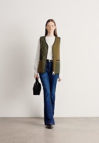 Πράσινο καπιτονέ γιλέκο με μαύρες λεπτομέρειες, φορεμένο πάνω από λευκό μακρυμάνικο τοπ. Συνδυασμένο με καμπανέ μπλε τζιν και κρατώντας μια μικρή μαύρη τσάντα.