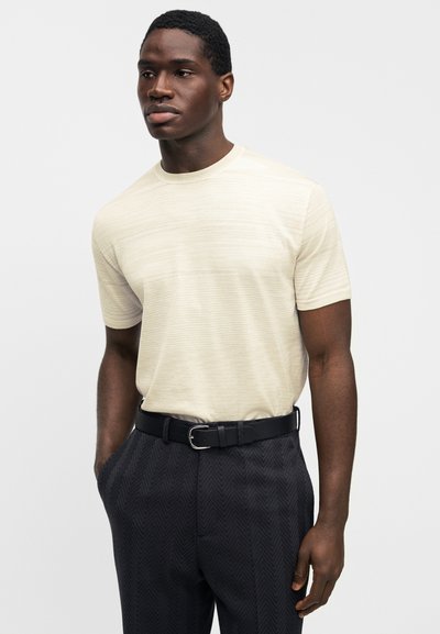 Κρεμ πουκάμισο με κοντά μανίκια και υφή, συνδυασμένο με σκούρα παντελόνια με ζιγκ-ζαγκ μοτίβο και μαύρη ζώνη, δημιουργώντας μια κομψή εμφάνιση.