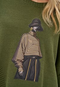 Sweter dziany w oliwkowym kolorze z dużą grafiką postaci w paskowanej bluzce i czarnych spodniach z złotymi akcentami.