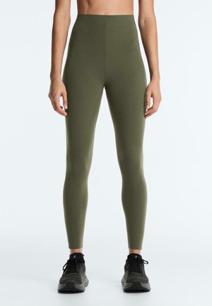 Slim fit olivovo zelené legíny nosené stojacou osobou v kombinácii s čiernymi športovými topánkami na jednoduchom pozadí.