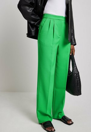 Femme portant un pantalon large vert vif, une veste en cuir noire, un sac à main noir tissé et des sandales ouvertes noires, debout contre un mur uni.