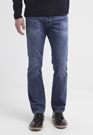 Mann trägt blaue Jeans, schwarze Schnürschuhe und ein schwarzes Langarmshirt, steht vor einem weißen Hintergrund.