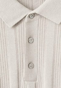 Camisa polo de punto beige con textura acanalada, tres botones grises y un diseño de cuello clásico.