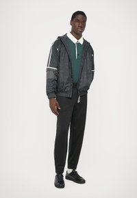 Jachetă neagră din nailon cu accente albe, tricou polo verde, pantaloni de trening negri și adidași negri, stând în fața unui fundal alb simplu.