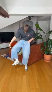 Un tânăr în interior ridică un picior, arătându-și blugii largi albastru deschis și adidașii albi într-un living cu plante și un televizor.