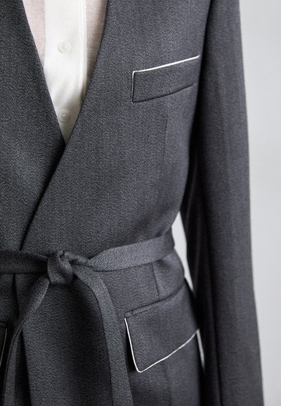 Szürke, texturált felületű blézer, fehér szegéllyel a zsebnél, megkötős anyagövvel és strukturált sziluettel.