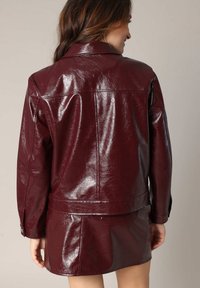 Veste en cuir synthétique bourgogne avec une finition brillante, dotée d'un col classique, de manches longues et d'un design ajusté, assortie à une mini-jupe coordonnée.