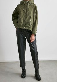 Grön huva-jacka med tryckknappar, två sidofickor och elastiska mudd, kombinerad med svarta, glänsande faux läderbyxor, bärs med svarta stövlar.