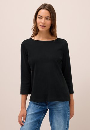 Donna che indossa un top lungo a maniche nere con scollatura a barca e jeans blu, in piedi contro uno sfondo beige chiaro.