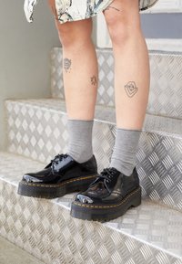 Scarpe con tacco platform in pelle verniciata nera, con punta arrotondata, suola testurizzata, cuciture gialle, indossate con calze grigie, mentre si trova su scale metalliche.