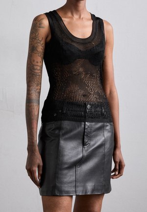 Vrouw draagt een mouwloos zwart mesh topje over een zwarte bh en een zwarte leren minirok, toont getatoeëerde linkerarm.