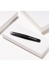 Pince à épiler noire pailletée avec une pointe effilée, portant le texte "TweezerMan", placée sur une boîte rectangulaire blanche.