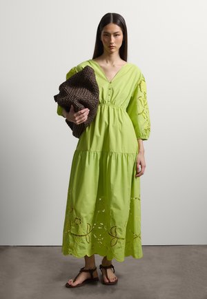 Donna con i capelli lunghi e scuri che indossa un vestito midi ricamato verde lime e sandali marroni, che tiene una grande borsa marrone a maglia grossa.