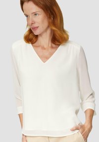 Blouse blanche à col en V et manches trois-quart, réalisée en tissu léger. Le design présente une silhouette épurée et un subtil éclat.
