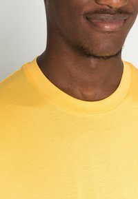Camiseta de algodón amarilla con escote redondeado y mangas cortas. Presenta una textura suave y costuras visibles a lo largo de los bordes.