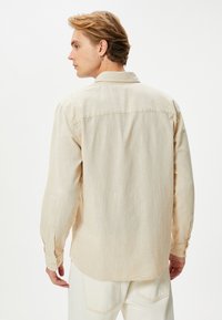 Lichtbeige linnen overhemd met lange mouwen, knoopmanchetten, subtiele textuur, rugpasdetail en een afgeronde zoom.