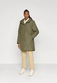 Γυναίκα που στέκεται σε χακί πράσινο μπουφάν με κουκούλα, μπεζ παντελόνια και λευκά αθλητικά παπούτσια, μπροστά από απλό ανοιχτό φόντο.