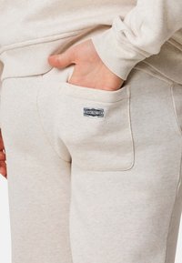 Ljust beige joggings med en mjuk textur, som har en sydd ficka och en liten "Denim Factory"-etikett på baksidan.