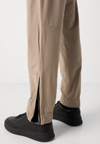 Bas des jambes portant un pantalon beige avec des fermetures éclair latérales ouvertes sur des baskets noires sur fond blanc.