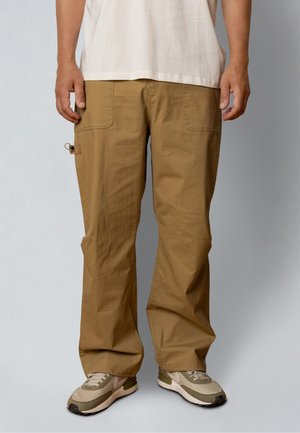 Man wearing beige wide-leg pants with front patch pockets and beige sneakers, standing against a neutral background.