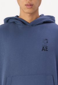 Μπλε ναυτικό φούτερ από βαμβάκι με μπροστινή τσέπη; διαθέτει γραφικό του Σνούπι και τα γράμματα "AE" σε μαύρο χρώμα στην αριστερή περιοχή του στήθους.