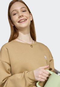 Brun sweatshirt med en vit logotyp, guldhjärt-halsband, håller en ljusgrön kanna med ett silverlock. Slät textur, avslappnad passform.