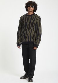 Pull en tricot avec un motif abstrait de rayures de tigre vertes et marine, associé à un pantalon noir à jambes larges et des chaussures noires.