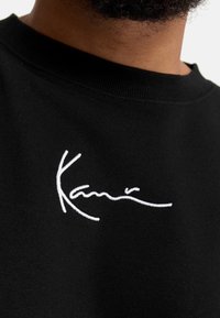 Sudadera negra con cuello redondo; cuenta con un texto bordado en blanco que dice "Kani". La tela parece suave con una textura lisa.