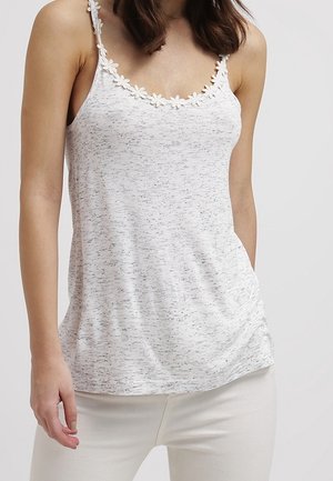 Femme portant un caraco blanc moucheté avec une bordure florale sur les bretelles et le décolleté, associé à un pantalon blanc, sur un fond uni.