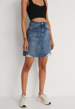 Frau trägt ein schwarzes, bauchfreies Top, einen blauen Jeansrock mit ausgefranstem Saum und weiße Turnschuhe, steht vor einem einfarbigen hellen Hintergrund.