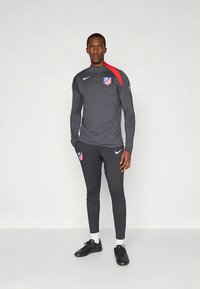 Chándal deportivo gris con mangas largas y pantalones, que presenta un acento rojo en el hombro y un logo. Zapatillas deportivas negras, textura lisa.