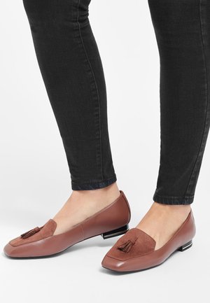 Mocassins à talons bas en cuir marron avec pompons portés avec un jean slim noir sur un fond clair.