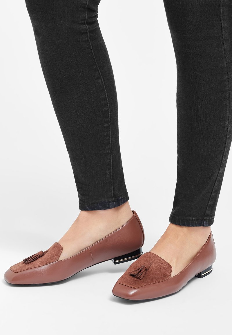 Mocassins à talons bas en cuir marron avec pompons portés avec un jean slim noir sur un fond clair.