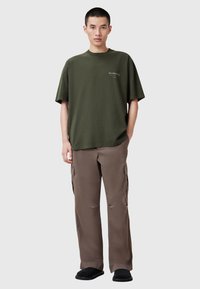 Camiseta de algodón verde oliva de manga corta y cuello redondo, combinada con pantalones cargo marrones que cuentan con bolsillos laterales y costuras visibles.