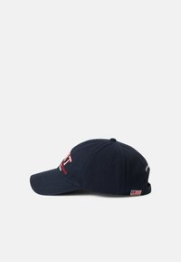 Navy blue cap made of cotton with a curved brim. Features a prominent logo in red and white on the front and a small flag accent on the side.