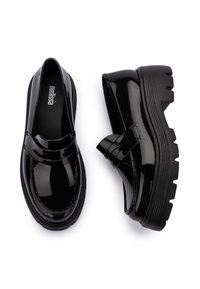 Mocasines de charol negro con suela de goma negra gruesa. Presentan un detalle de lazo plano y costura a lo largo del borde superior.