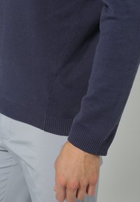 Navy blue knitted sweater with ribbed cuffs and hem, featuring a smooth texture and fitted design, shown alongside light-colored pants.