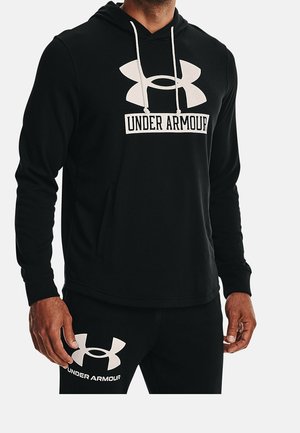 Mann trägt einen schwarzen Under Armour Hoodie und passende schwarze Hose mit großen weißen Under Armour Logos auf beiden.