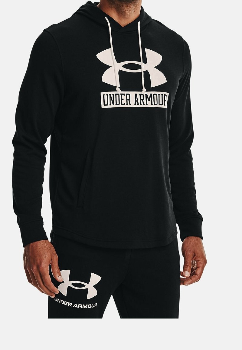 Homme portant un sweat à capuche noir Under Armour et un pantalon noir assorti avec de grands logos blancs Under Armour sur les deux.