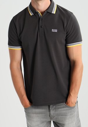 Mann trägt ein schwarzes Hugo Boss Polohemd mit gelbem und grauem Besatz an Kragen und Ärmeln, Hände in den Taschen, hellgraue Jeans.